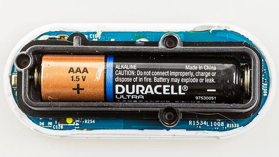 An opened oblong electronic device reveals a blue circuit board surrounding a black battery holder containing one Duracell Ultra AAA 1.5-volt battery.