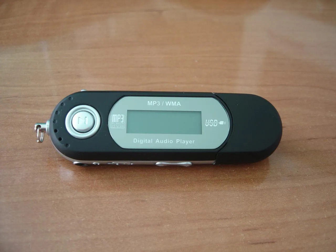 Black and silver portable digital audio player on a wooden surface, with a blank rectangular screen, circular play button, USB cap and metal keyring loop.