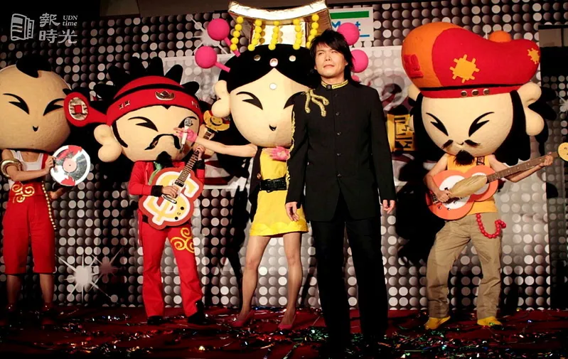 A performer stands between large Good Gods character mascots on a promotional stage in Taiwan.